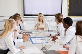 Female High School Teacher Sitting At Table With Teenage Pupils Wearing Uniform Teaching Lesson