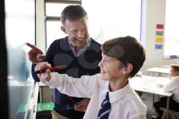 High School Teacher With Male Student Wearing Uniform Using Interactive Whiteboard During Lesson