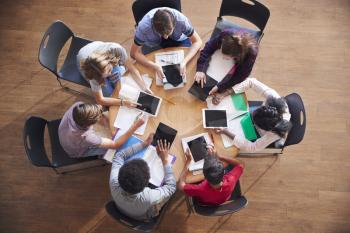 Overhead Shot Of High School Pupils Using Digital Tablets In Group Study Around Tables