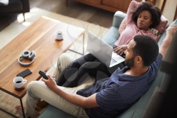 Couple Sitting On Sofa At Home Using Laptop Computer And Watching TV