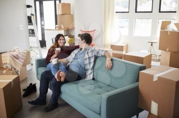 Happy Couple Resting On Sofa Surrounded By Boxes In New Home On Moving Day