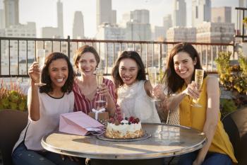 Portrait Of Female Friends Making Toast To Celebrate Birthday On Rooftop Terrace With City Skyline In Background