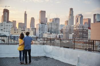 Rear View Of Couple On Rooftop Terrace Looking Out Over City Skyline At Sunset