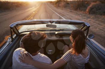 Rear View Of Couple On Road Trip Driving Classic Convertible Car Towards Sunset