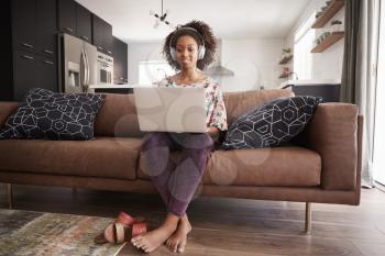 Woman Wearing Wireless Headphones Sitting On Sofa At Home Using Laptop