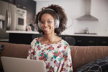 Portrait Of Woman Wearing Wireless Headphones Sitting On Sofa At Home Using Laptop