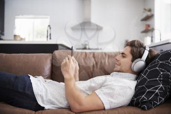 Man Wearing Wireless Headphones Lying On Sofa At Home Streaming From Mobile Phone