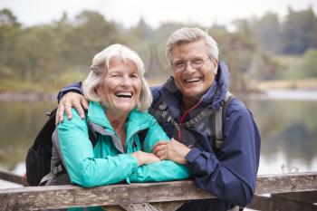 Happy senior couple leaning on a wooden fence laughing to camera, close up, Lake District, UK