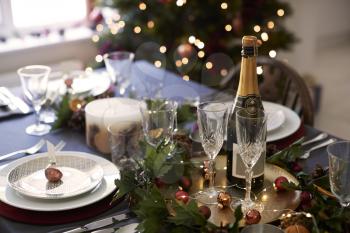 Christmas table setting with glasses and a bottle of champagne, bauble name card holder arranged on a plate and green and red table decorations
