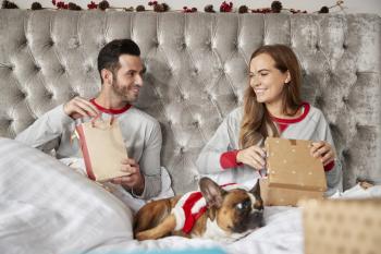 Couple In Bed At Home With Dog Dressed In Santa Costume Opening Gifts On Christmas Day