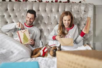 Couple In Bed At Home With Dog Dressed In Santa Costume Opening Gifts On Christmas Day