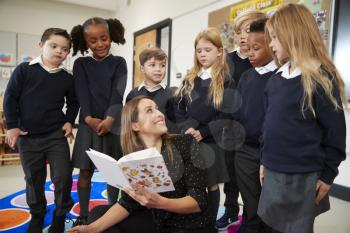 Primary school teacher reading a book in class turning around to look at the kids standing behind her, front view