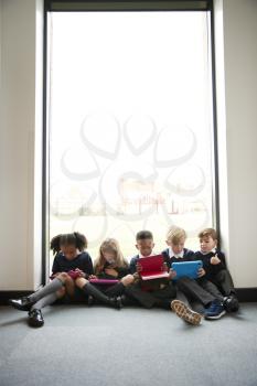 Primary school kids sitting together on the floor in front of a window in a school corridor using tablet computers, vertical