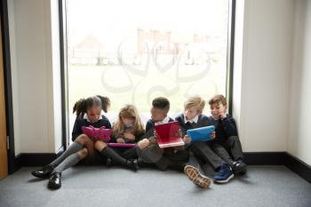 Five primary school kids sitting in a row on the floor in front of a window in a school corridor looking at tablet computers, front view, close up