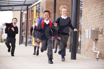 Happy primary school kids, wearing school uniforms and backpacks, running on a walkway outside their school building, front view, close up