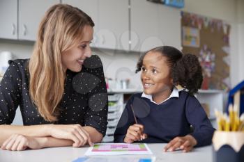 Young female primary school teacher working one on one with a schoolgirl at a table in a classroom, both looking at each other smiling, close up