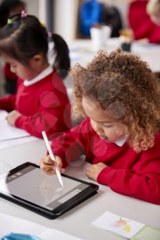 Elevated view of young schoolgirl wearing school uniform sitting at desk in an infant school classroom using a tablet computer and stylus, close up, vertical