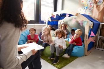Female infant school teacher sitting on a chair reading a book to a group of children sitting on bean bags in a comfortable corner of the classroom, elevated view, close up