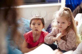 Two infant school girls sitting on bean bags in a comfortable corner of the classroom listening to their teacher reading a story, view over teachers shoulder