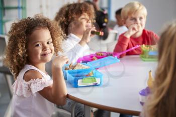Close up of smiling young children sitting at a table eating their packed lunches together at infant school, girl smiling to camera, selective focus