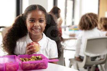 Young black schoolgirl sitting at a table smiling and holding an apple in a kindergarten classroom during her lunch break, close up