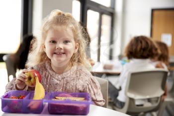 Young white schoolgirl sitting at a table smiling and holding an apple in a kindergarten classroom during her lunch break, close up