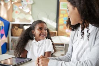 Young black schoolgirl sitting at a table with a tablet computer in an infant school classroom learning one on one with female teacher, smiling at each other, close up