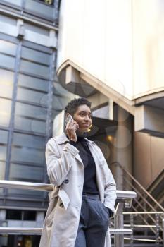 Fashionable young black businesswoman standing against handrail in the city talking on smartphone, vertical