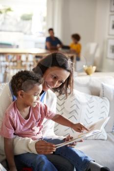 Close up of young mother sitting on a sofa in the living room reading a book with her toddler son, sitting on her knee, father and daughter sitting at a table in the background, vertical