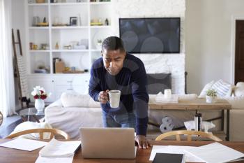 Millennial Hispanic man stands leaning on table in the dining room holding cup and looking down at his laptop computer screen