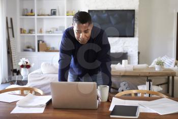 Millennial Hispanic man stands leaning on table in the dining room looking down at his laptop computer screen