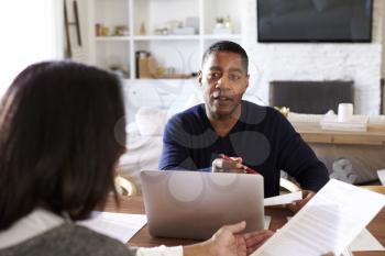 Millennial man with laptop computer giving financial advice to a woman sitting at the table holding a document in her dining room, close up, selective focus