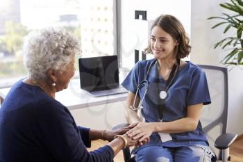 Nurse Wearing Scrubs In Office Reassuring Senior Female Patient And Holding Her Hands