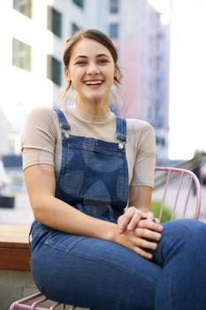 Trendy young Hispanic woman wearing dungarees sitting in the street laughing to camera, close up