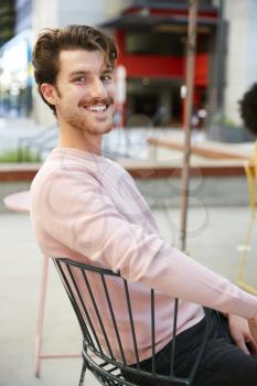Millennial white hipster man sitting outside a cafe in the street, turns smiling to camera, close up