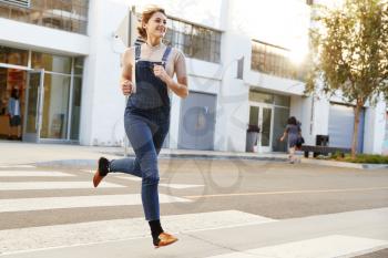 Fashionable young Hispanic woman in dungarees running across the street, full length, close up