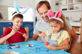 Father With Children Wearing Rabbit Ears Decorating Easter Eggs At Home Together