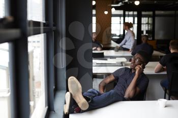 Young black male creative sitting in an office with his feet up on the desk using phone, close up