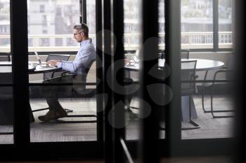 Middle aged white businessman working alone in a meeting room, seen through glass door