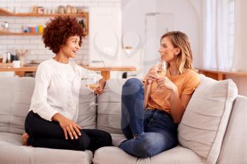 Two Female Friends Relaxing On Sofa At Home With Glass Of Wine Talking Together