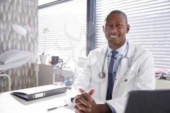 Portrait Of Smiling Male Doctor Wearing White Coat With Stethoscope Sitting Behind Desk In Office