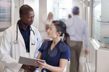 Doctor And Nurse Consulting Over Digital Tablet In Busy Hospital Corridor
