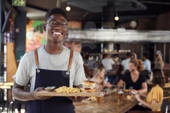 Portrait Of Waiter Serving Food To Customers In Busy Bar Restaurant