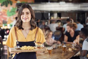 Portrait Of Waitress Serving Food To Customers In Busy Bar Restaurant