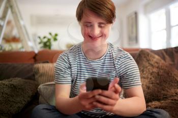 Young Downs Syndrome Man Sitting On Sofa Using Mobile Phone At Home