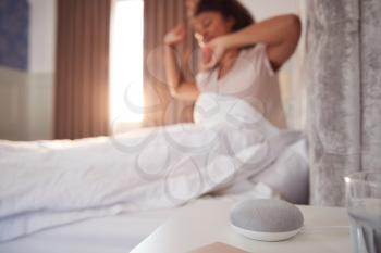 Woman Waking Up In Bed With Voice Assistant On Bedside Table Next To Her