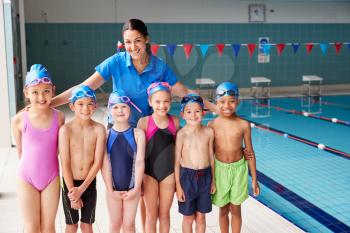 Portrait Of Female Coach With Children In Swimming Class Standing Edge Of Indoor Pool