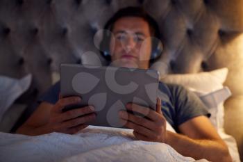 Man Wearing Wireless Headphones Lying In Bed Watching Digital Tablet Before Going To Sleep