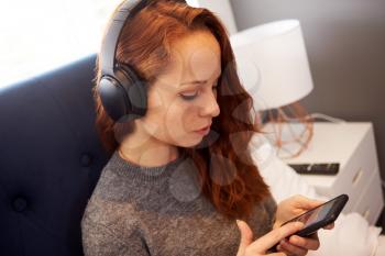 Female College Student Wearing Headphones Sitting On Bed In Shared House With Mobile Phone