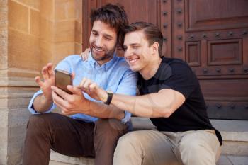 Male Gay Couple On Vacation Sitting Outdoors On Steps Of Building Looking At Mobile Phone